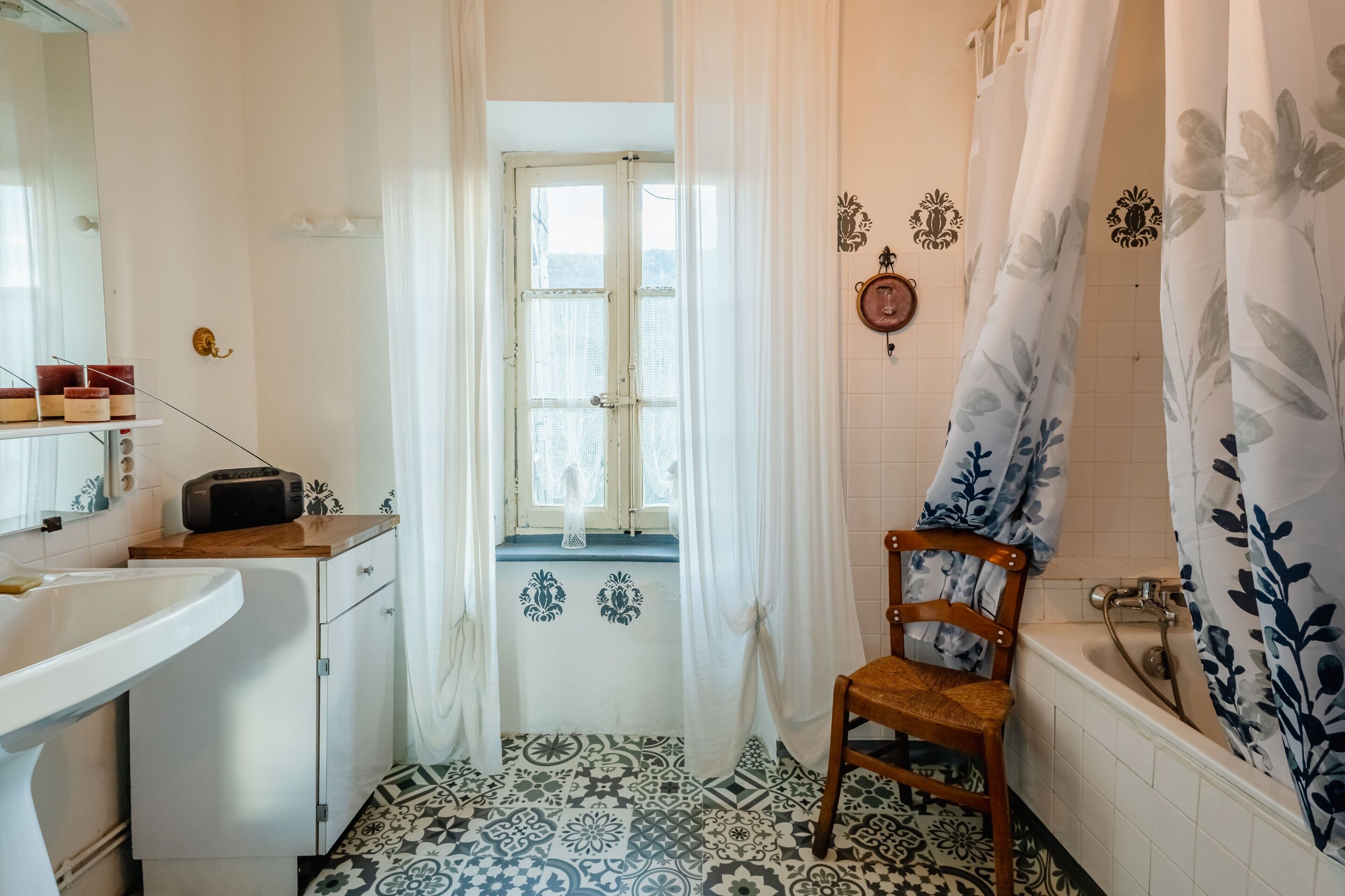 Une salle de bain avec une baignoire, une chaise en bois, un lavabo, une radio et une fenêtre avec des rideaux.