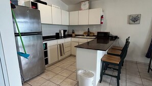 Una cocina con armarios blancos, un refrigerador de acero inoxidable, un lavamanos y un pequeño comedor con dos taburetes de madera.