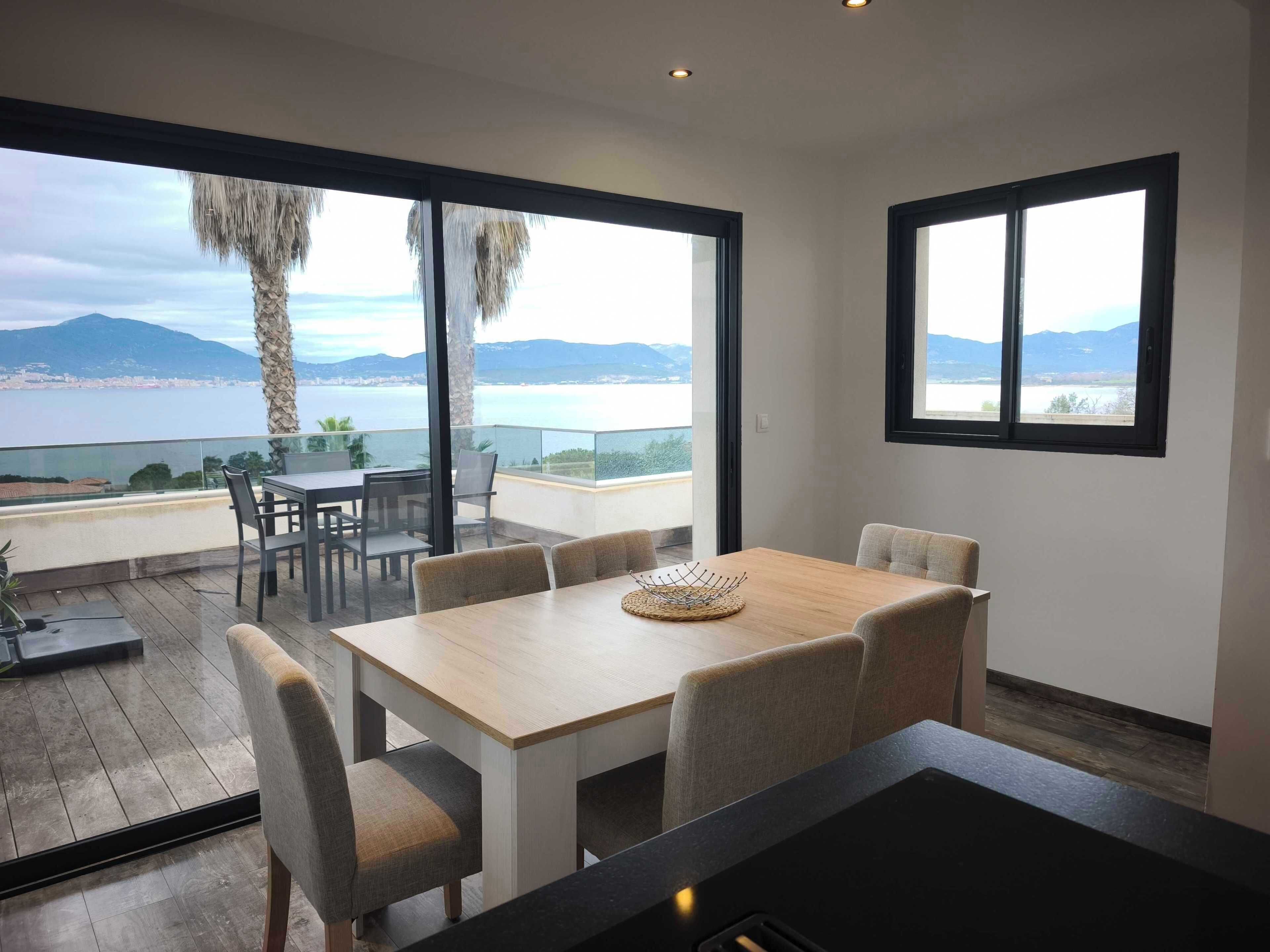 Een moderne eethoek met een houten tafel en stoelen, grote ramen met uitzicht op zee en bergen, en een glazen balustrade.