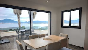 A modern dining area with a wooden table and chairs, large windows with a view of the sea and mountains, and a glass railing. - A serene stay with a sea view (Grosseto-Prugna)