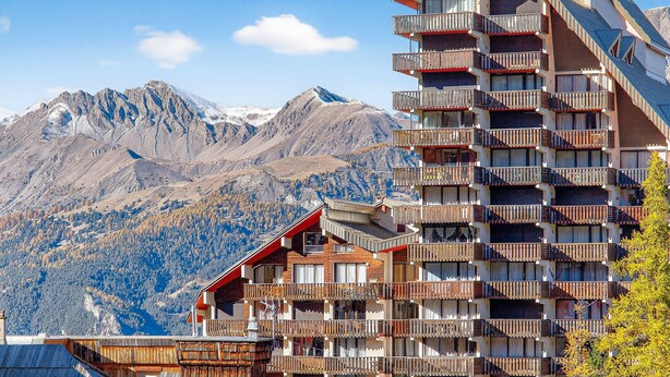 A multi-story hotel with balconies, a mountain backdrop, and a ski slope visible in the distance.