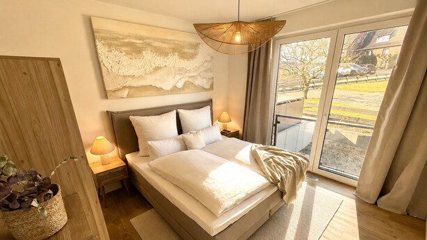 A bedroom with a bed, a nightstand, a lamp, a large window with a view, and a textured wall piece. - Pier 2: 200m to the sea, terrace, sauna, fireplace (first floor) (Scharbeutz)
