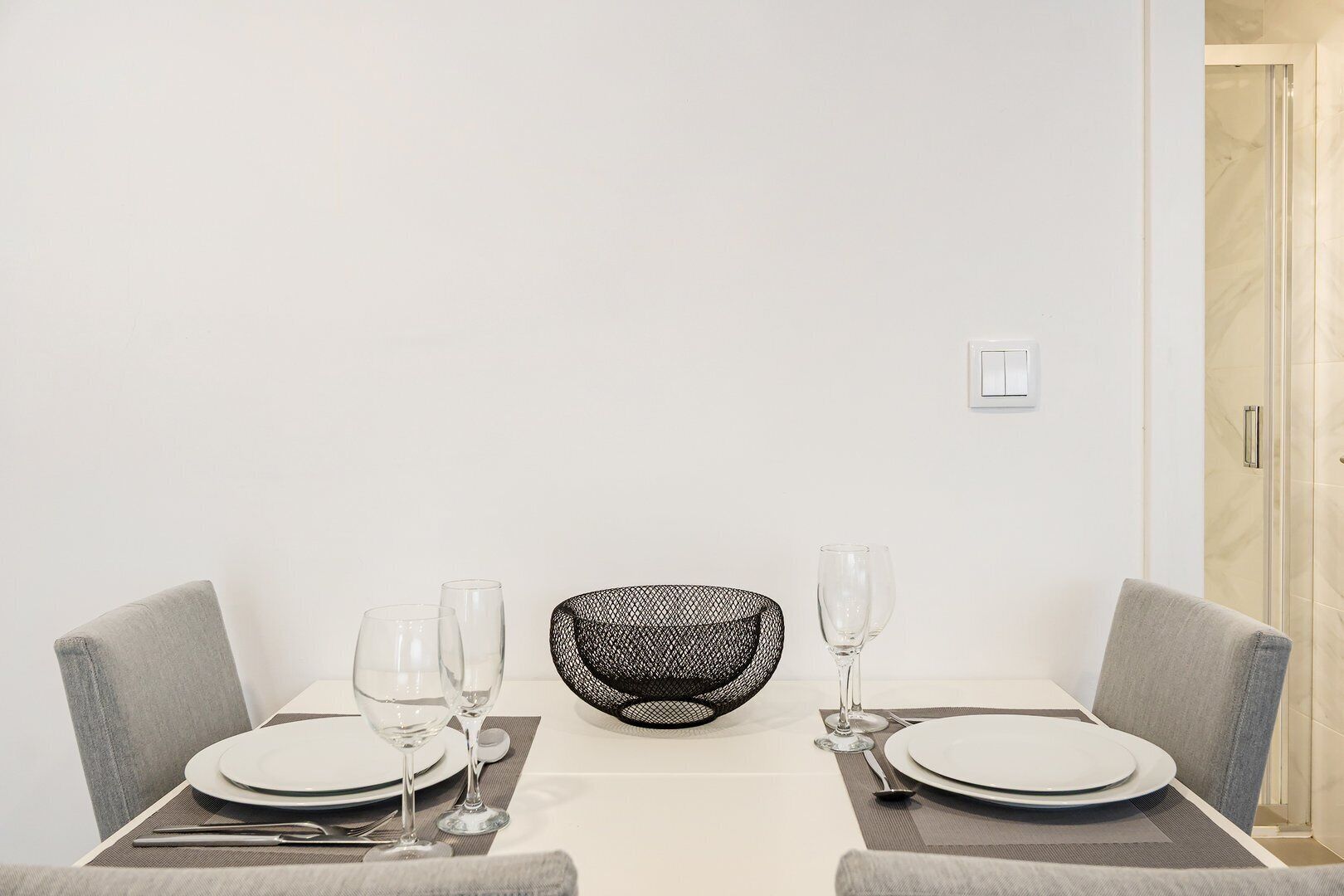 A dining table set with plates, glasses, and cutlery, with a mesh bowl and a small white object on the table.