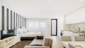 A modern, well-lit hotel room with a bed, a TV, a sofa, a dining table, and a kitchen area.