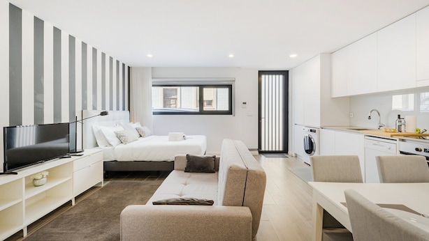 A modern, well-lit hotel room with a bed, a TV, a sofa, a dining table, and a kitchen area with a washing machine.