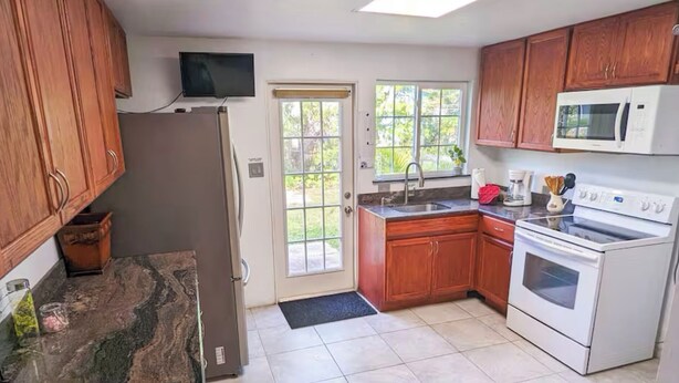 A kitchen with wooden cabinets, a refrigerator, a microwave, a stove, and a window with a view of greenery.