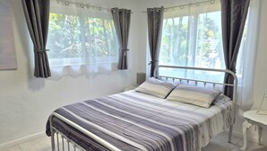 A bedroom with a bed, curtains, a bedside table, and a view of greenery outside.