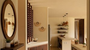 Un pasillo con suelo de madera, un espejo, una planta y una zona de cocina con estantes y luces colgantes.