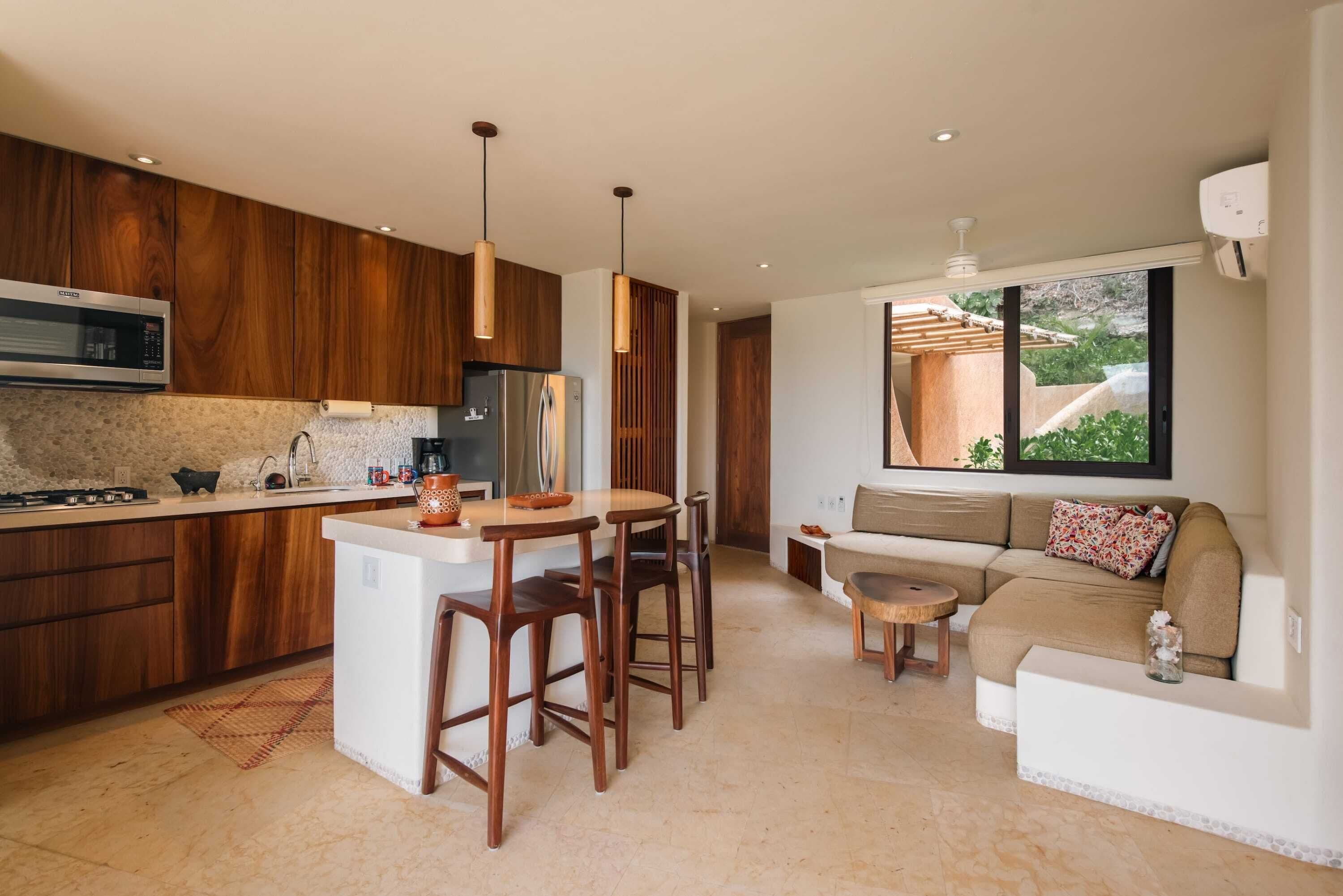 A modern kitchen with wooden cabinets, a central island, and pendant lighting. There is a dining area with a sofa and a small table.