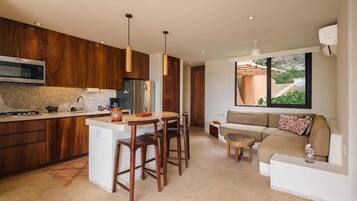 A modern kitchen with wooden cabinets, a central island, and pendant lighting. There is a dining area with a sofa and a small table.
