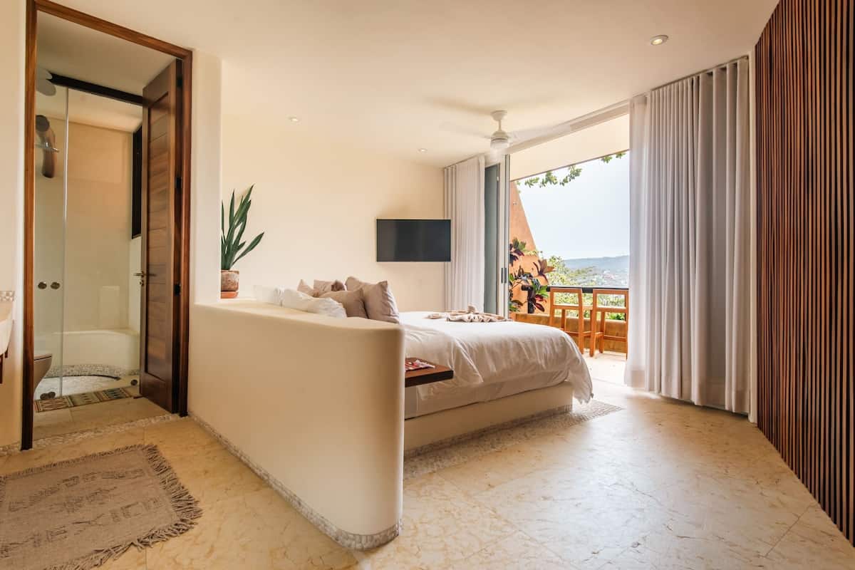 Ein modernes Schlafzimmer mit Bett, einer Couch, einem Fernseher und einem Balkon mit Vorhängen.