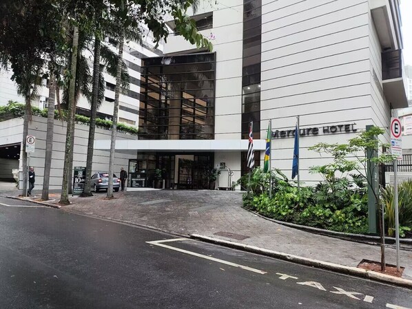 A modern hotel entrance with a glass facade, a parking area, and a pedestrian pathway.