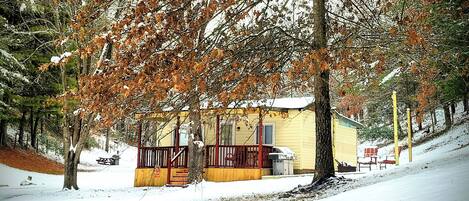 A small yellow cabin with a red porch, surrounded by snow-covered trees and a snowy landscape.