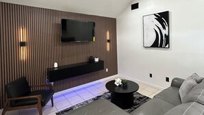 A modern living room with a flat-screen TV mounted on a wooden panel wall, a ceiling fan, a black coffee table, and a grey sofa.