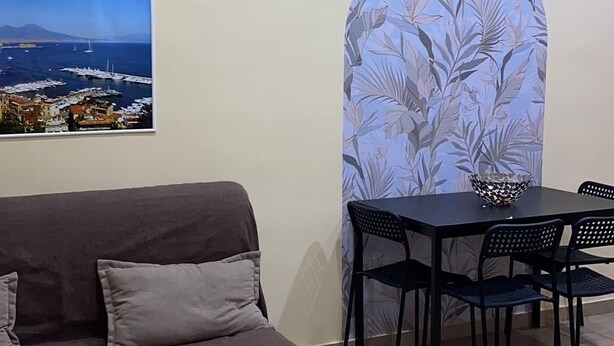 Ein Wohnzimmer mit braunem Sofa, einem Esstisch mit vier Stühlen, einem Wandbild mit Küstenlandschaft und einer Wandverkleidung mit Muster.