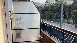 Ein Balkon mit rundem Tisch und zwei Stühlen, einem großen Wasserspender und Blick auf eine Stadtlandschaft mit Gebäuden und einer roten Kuppelstruktur.