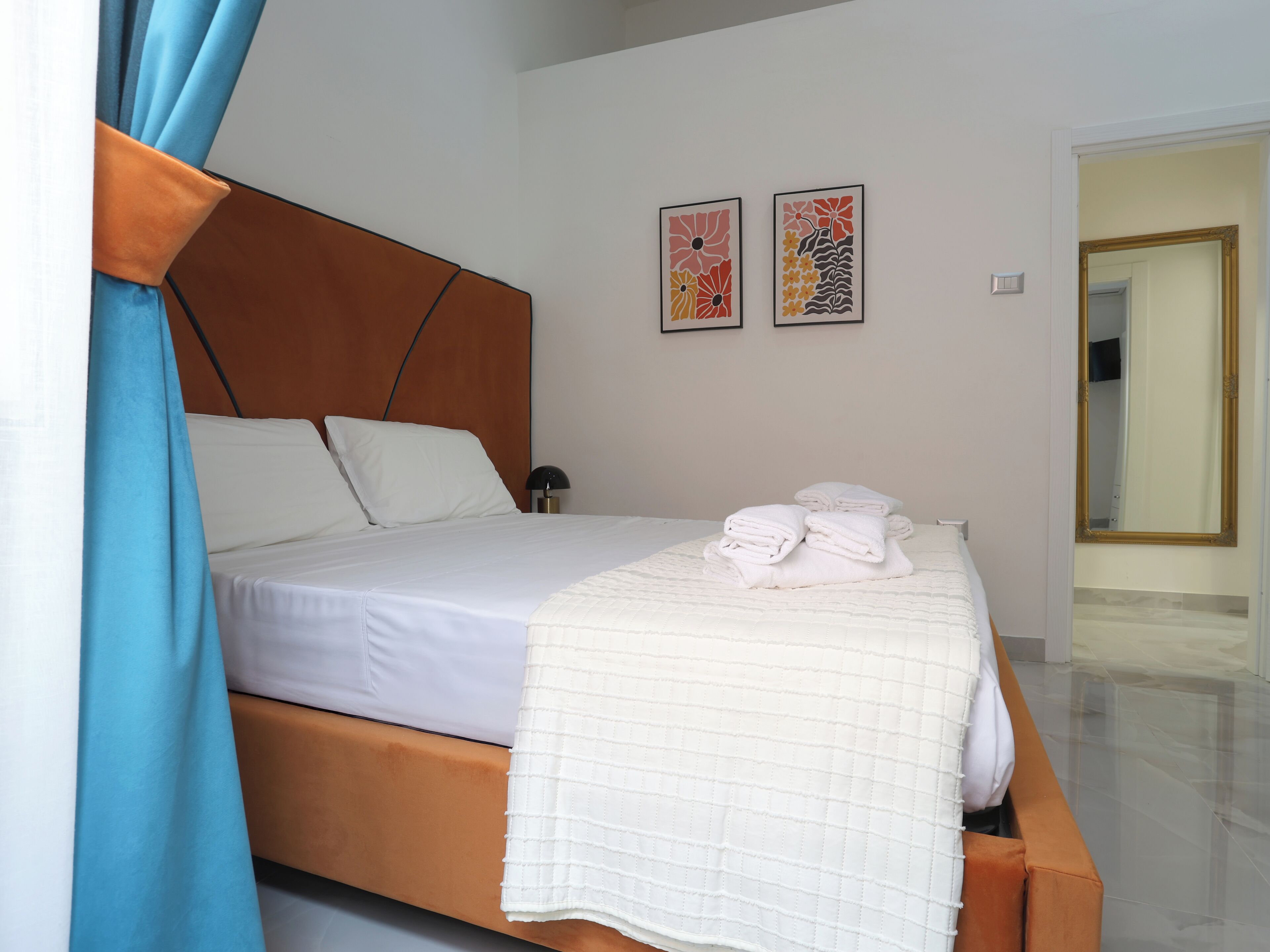 A neatly made bed with white linens and a quilted blanket, a bedside lamp, and two framed artworks on the wall.