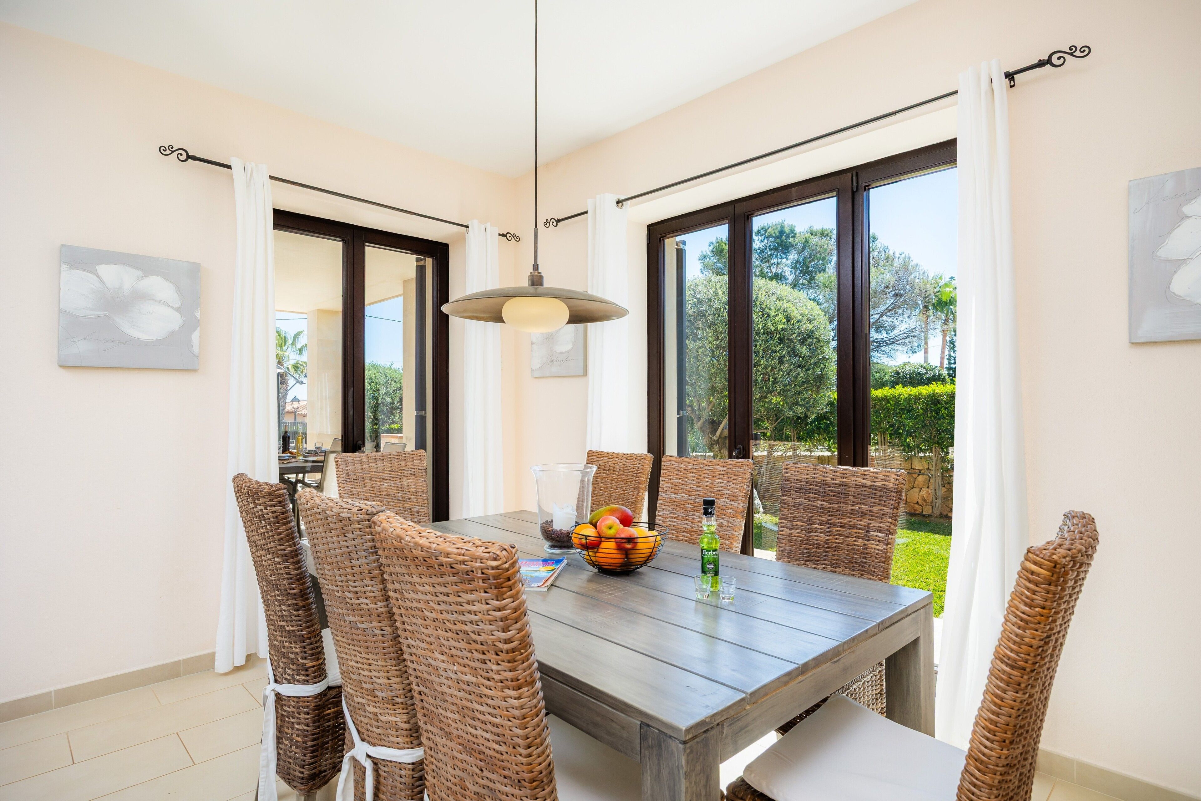 A dining area with wicker chairs, a wooden table, a fruit bowl, and a bottle of water.