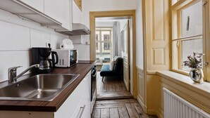 A narrow kitchen with a wooden countertop, stainless steel sink, and a coffee maker. A hallway leads to a living area with a sofa and a window with a vase of flowers. - Charming Apartment in Old Town (Stockholm)
