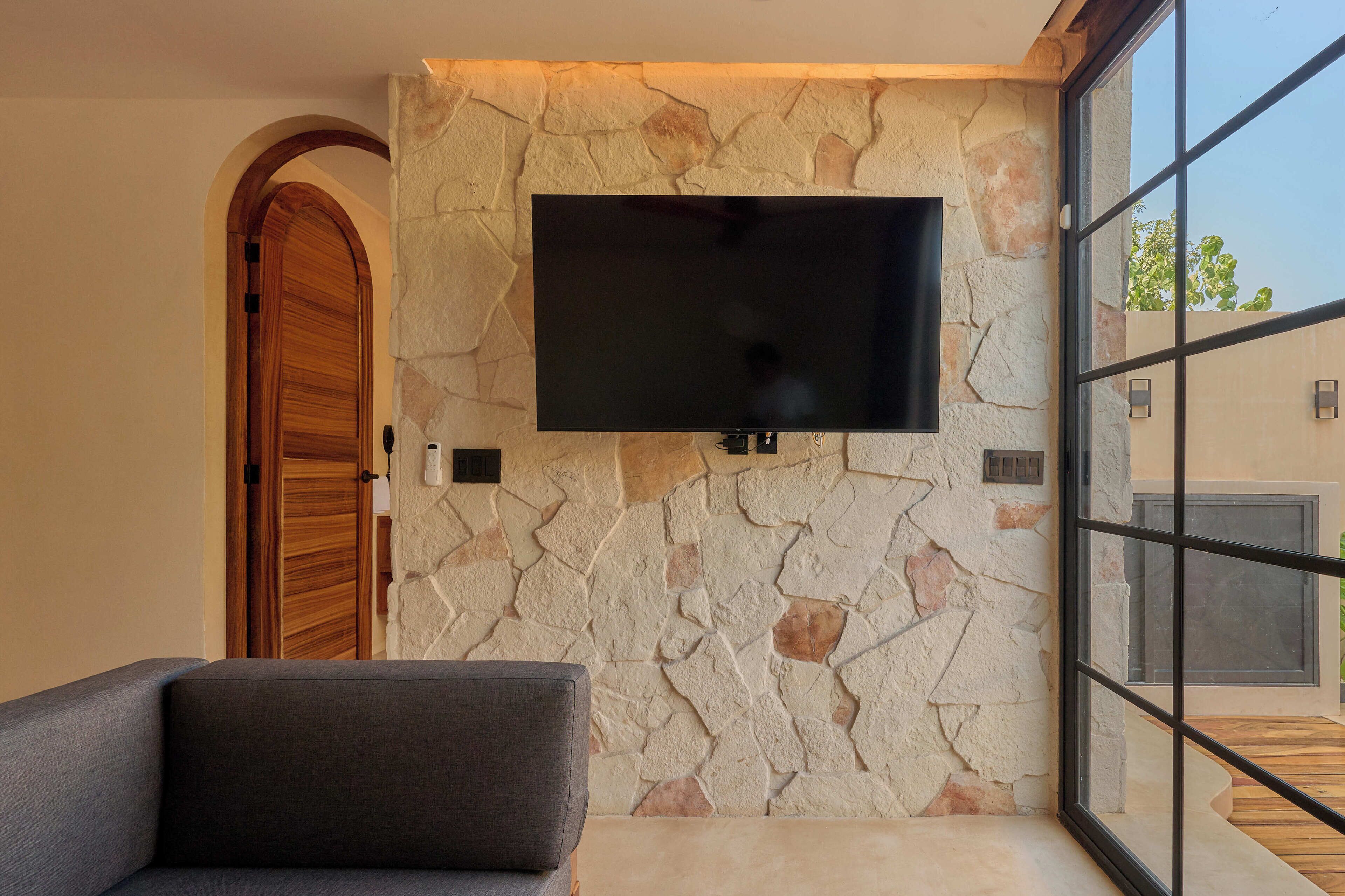 A modern interior with a stone wall, a flat-screen TV mounted above it, a wooden door, and a large window.