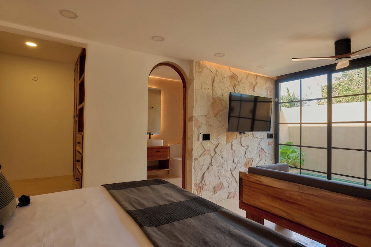 A bedroom with a bed, a wooden bench, a TV mounted on the wall, and a stone-textured wall.