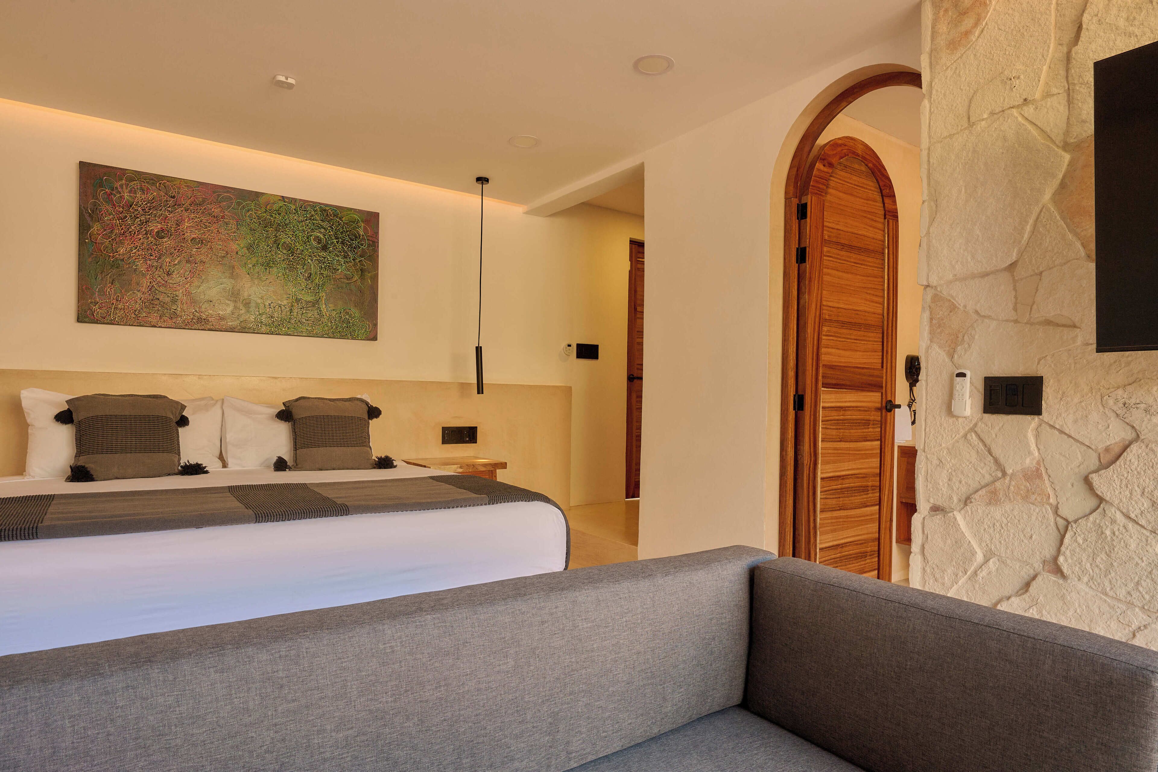 A modern bedroom with a large bed, a gray sofa, a stone wall, and a wooden door.