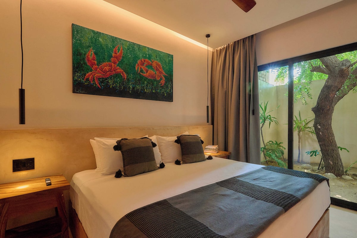 A hotel room with a bed, a painting of crabs, a window with a view of a tree, and a nightstand with a lamp.