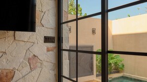 A room with a stone wall, a flat-screen TV, and a view of an outdoor swimming pool through a large window. - Hotel Compatir (Santa María Colotepec)