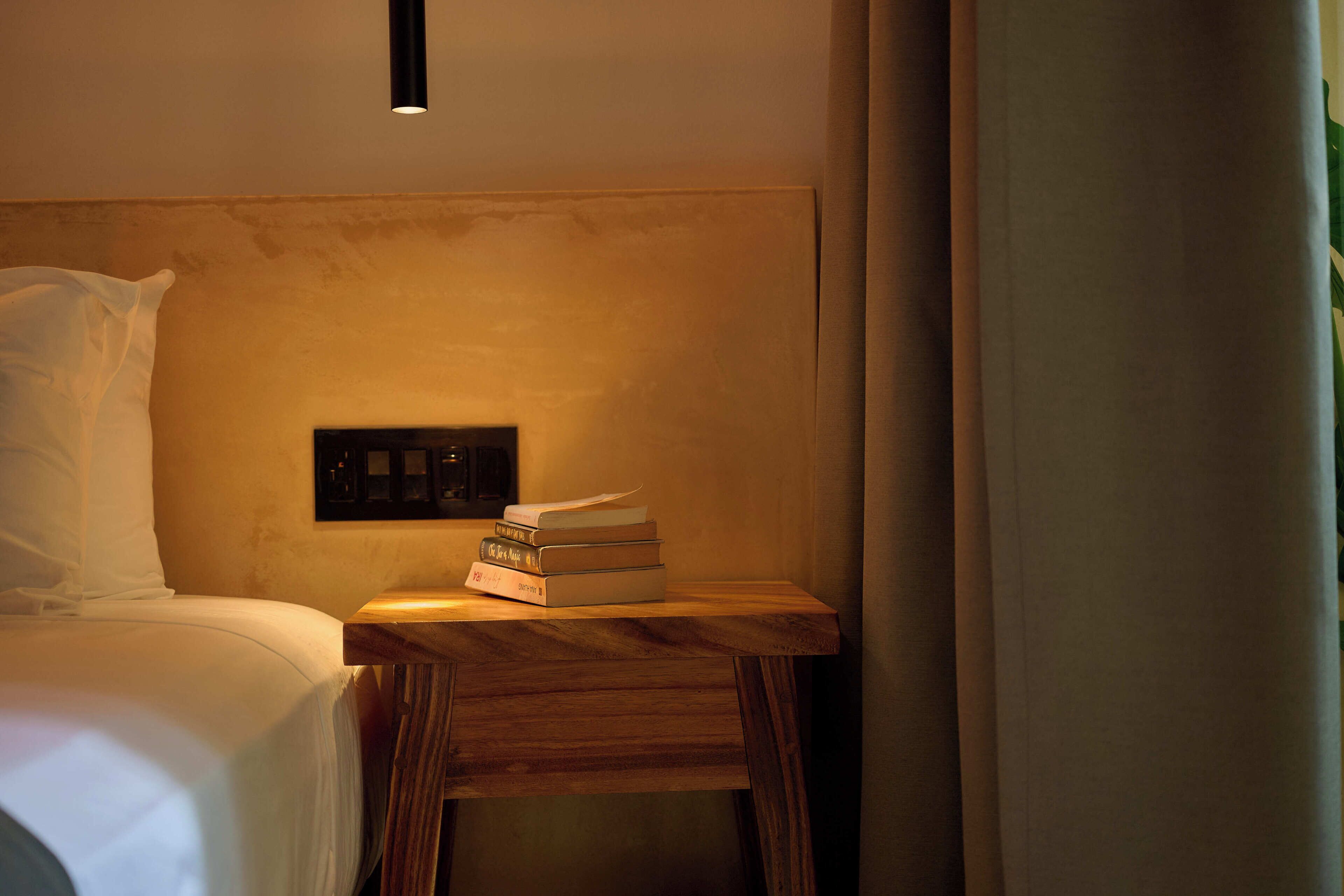 A neatly made bed with white linens, a small wooden nightstand with a stack of books, and a wall-mounted light fixture.