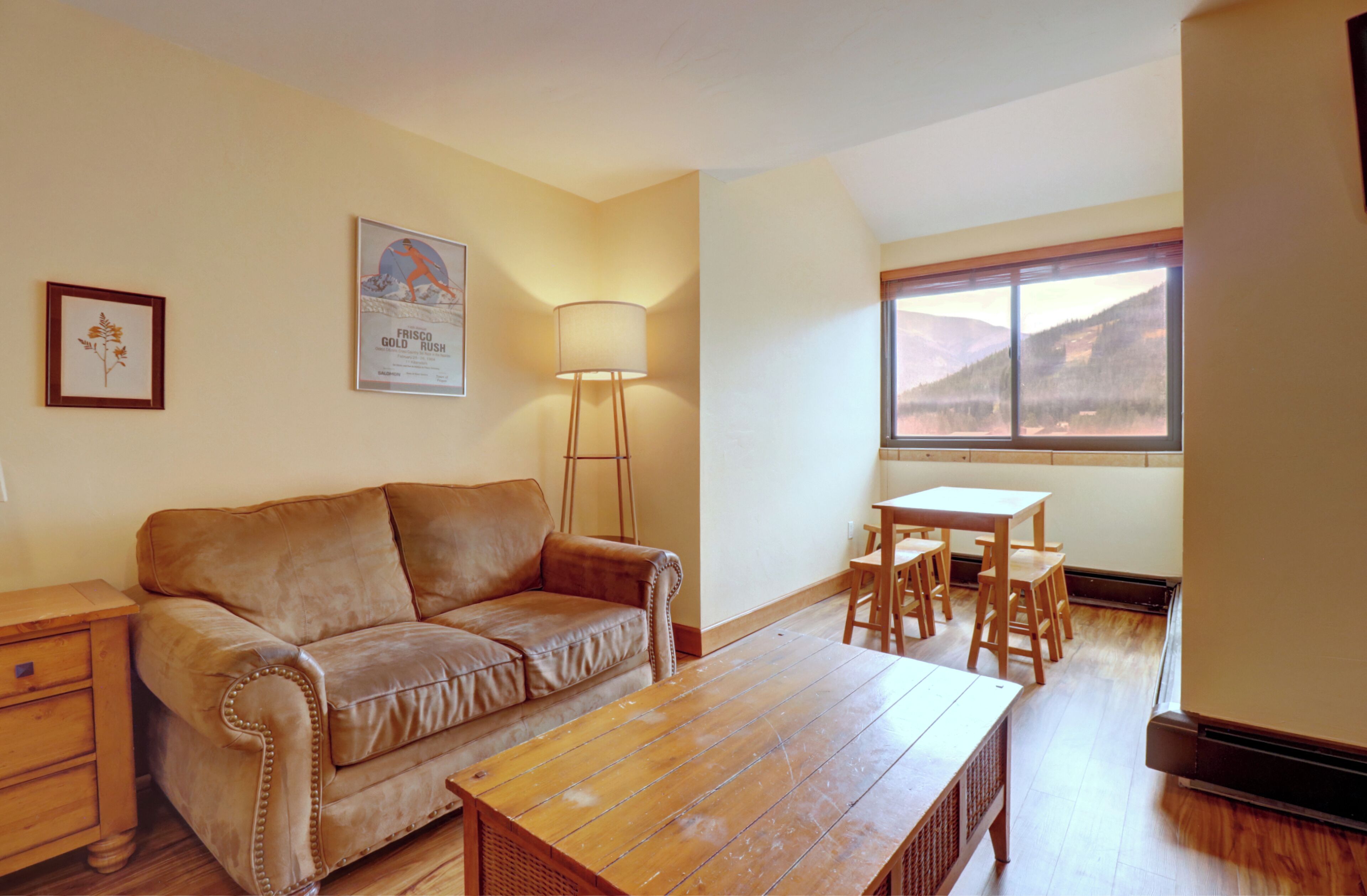 A living room with a brown leather sofa, a wooden coffee table, a lamp, a framed picture, and a window with a mountain view.