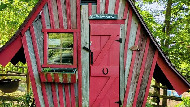 Un edificio rústico de madera con una puerta roja y un ventanal que indica “Baños”.
