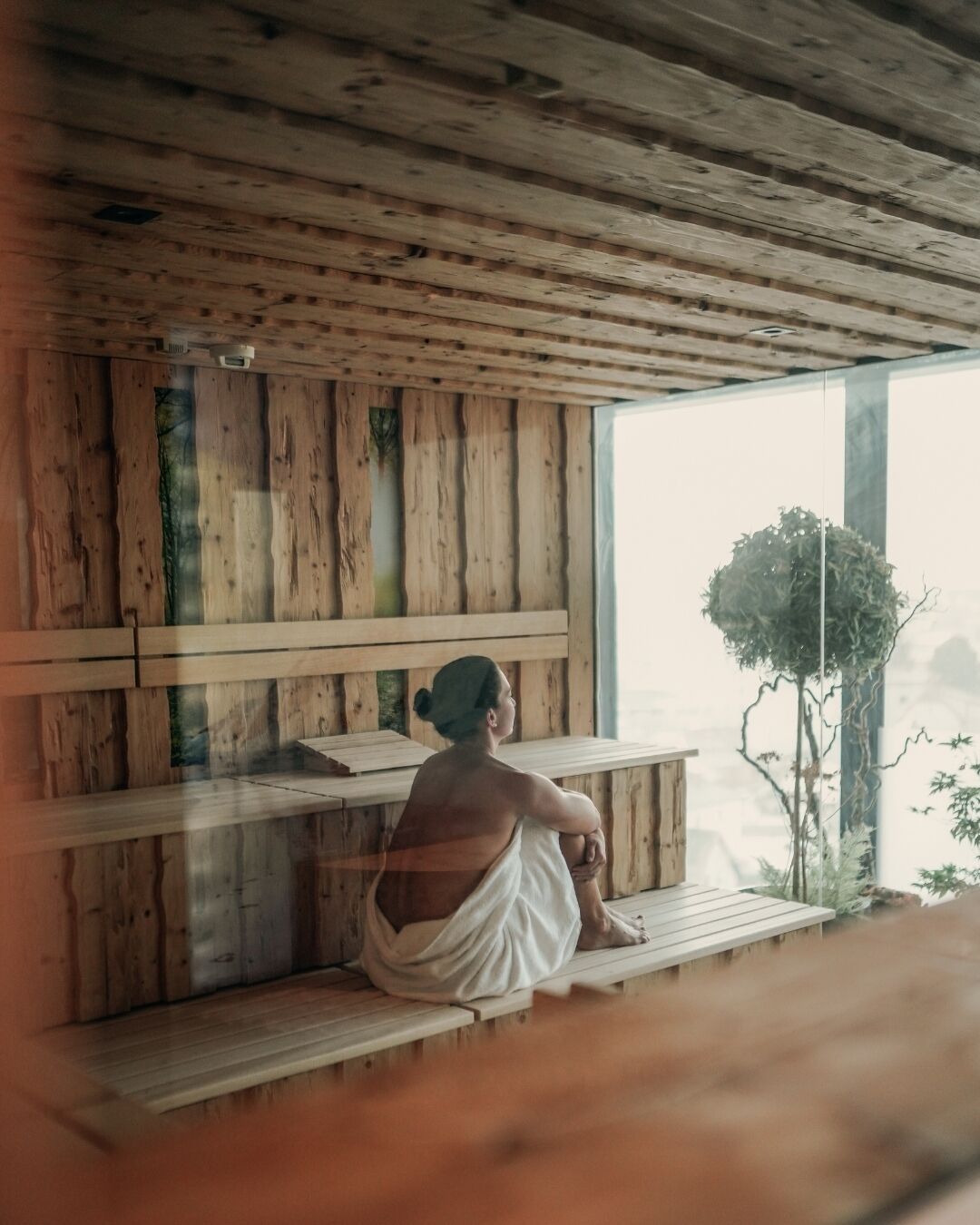 A person sitting on a wooden bench in a room with wooden walls and a large window.