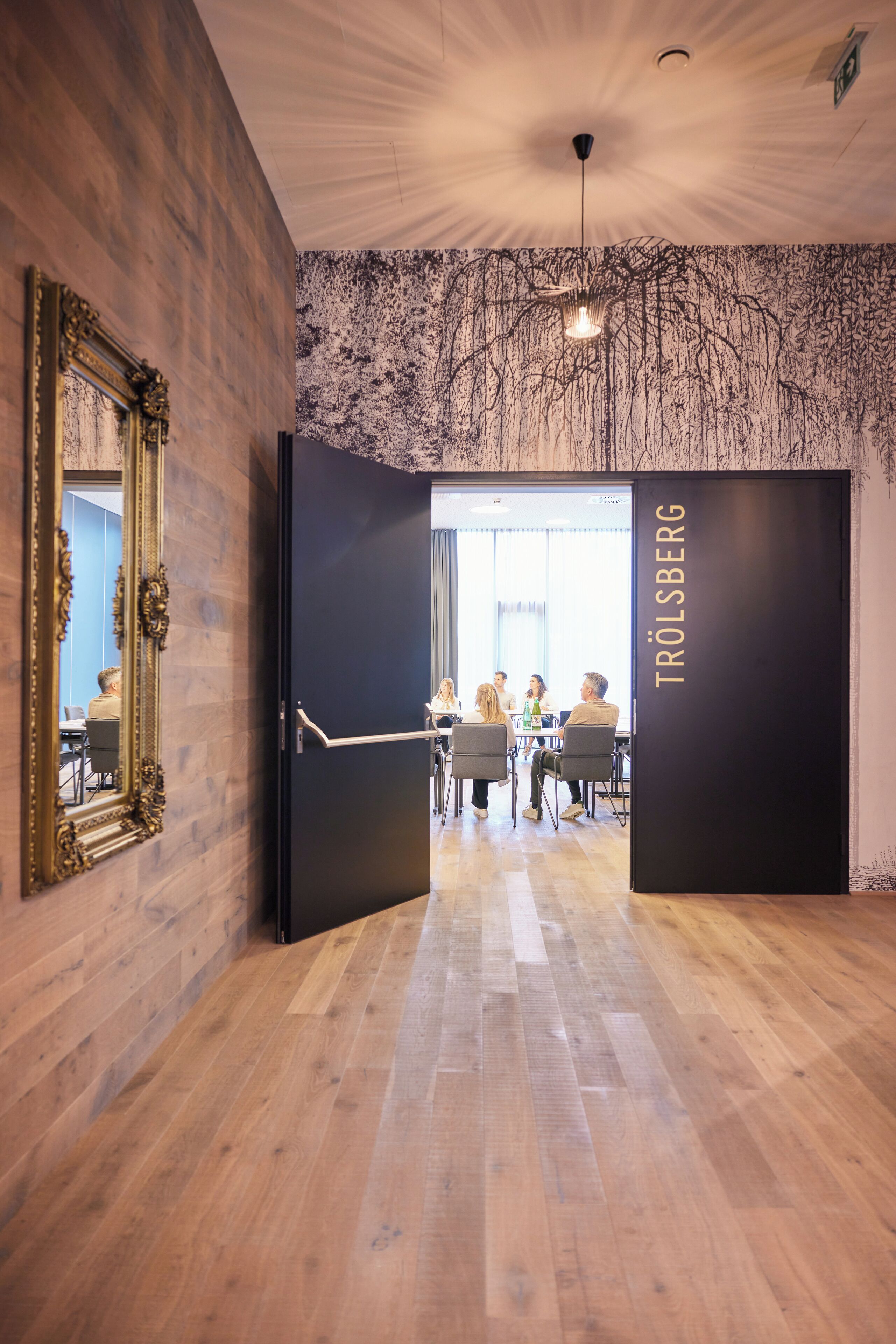 A conference room with a wooden floor, a large mirror, and a door leading to a meeting area with tables and chairs.