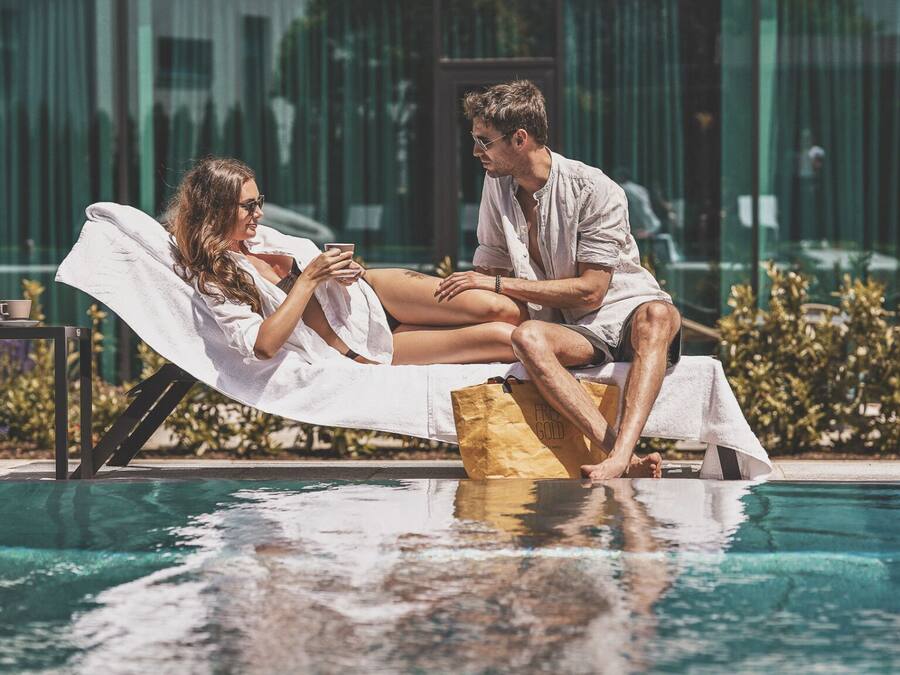 A couple relaxing on a lounge chair by a pool with a modern building in the background.