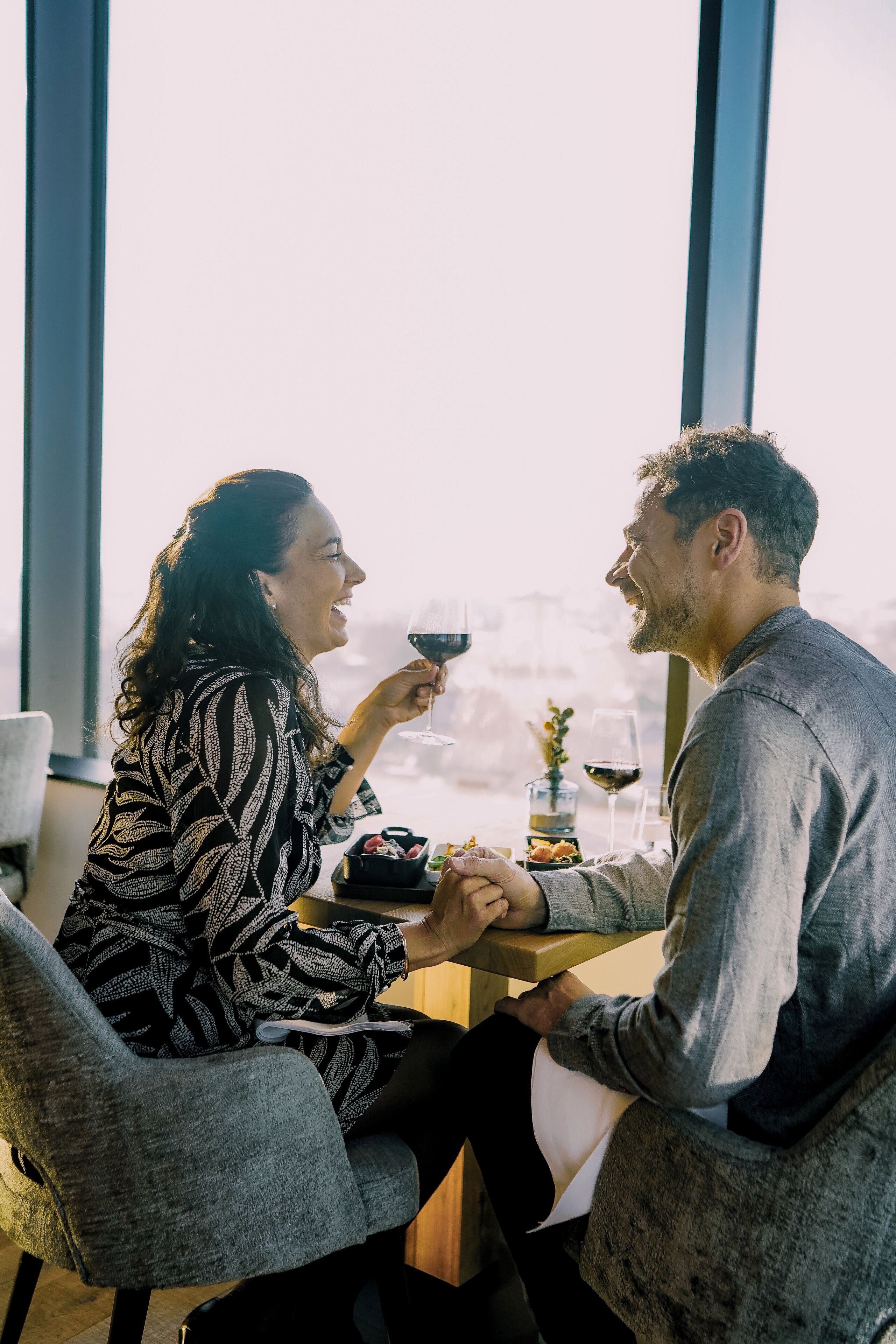 Two people sitting at a table with wine glasses, a small plant, and a bowl of fruit.