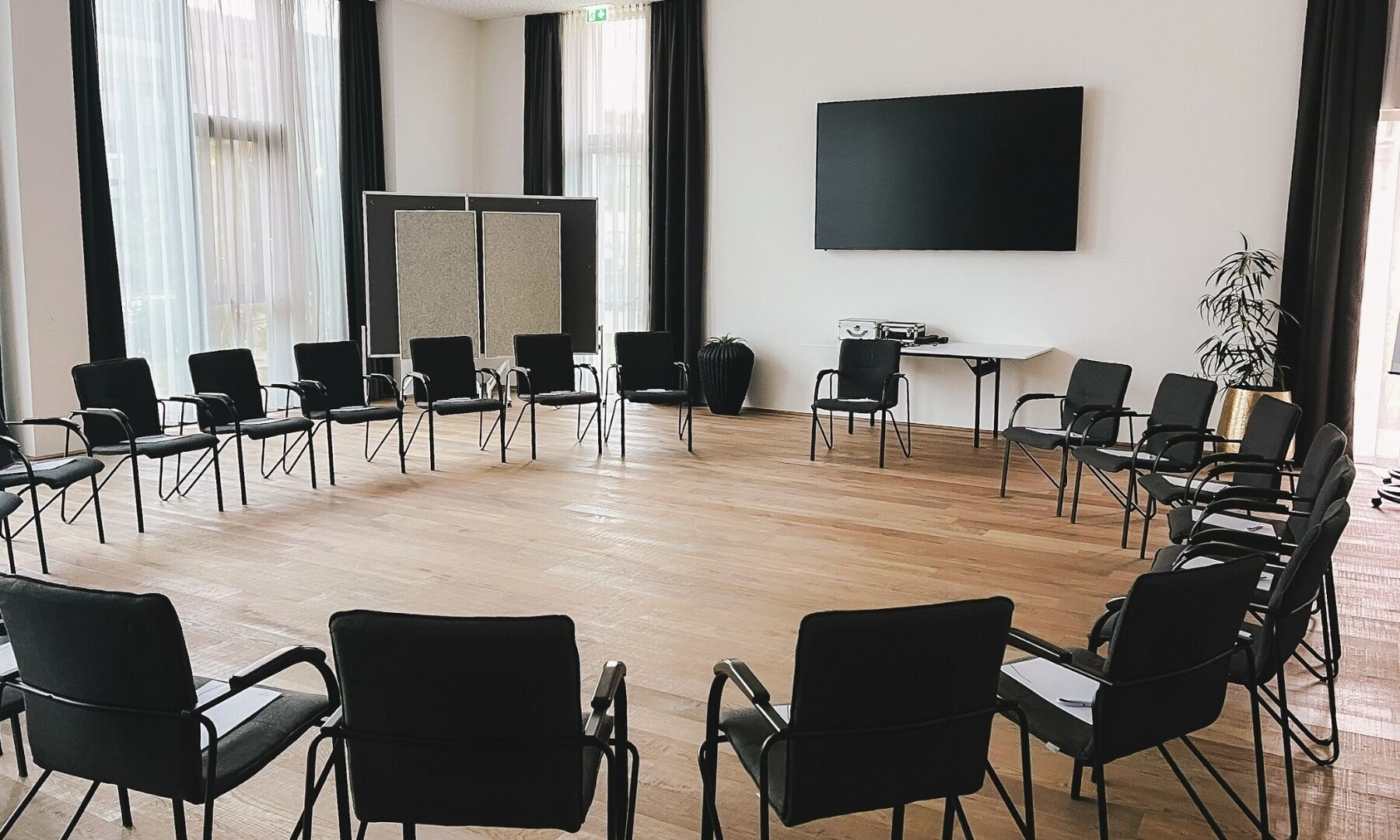 Een vergaderruimte met een groot zwart scherm aan de muur, zwarte stoelen in een halve cirkel gerangschikt, en een houten vloer.