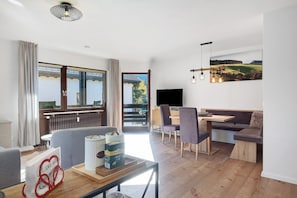 Uma sala de estar moderna com sofá, mesa de jantar e cadeiras, uma janela grande com cortinas e um quadro na parede.