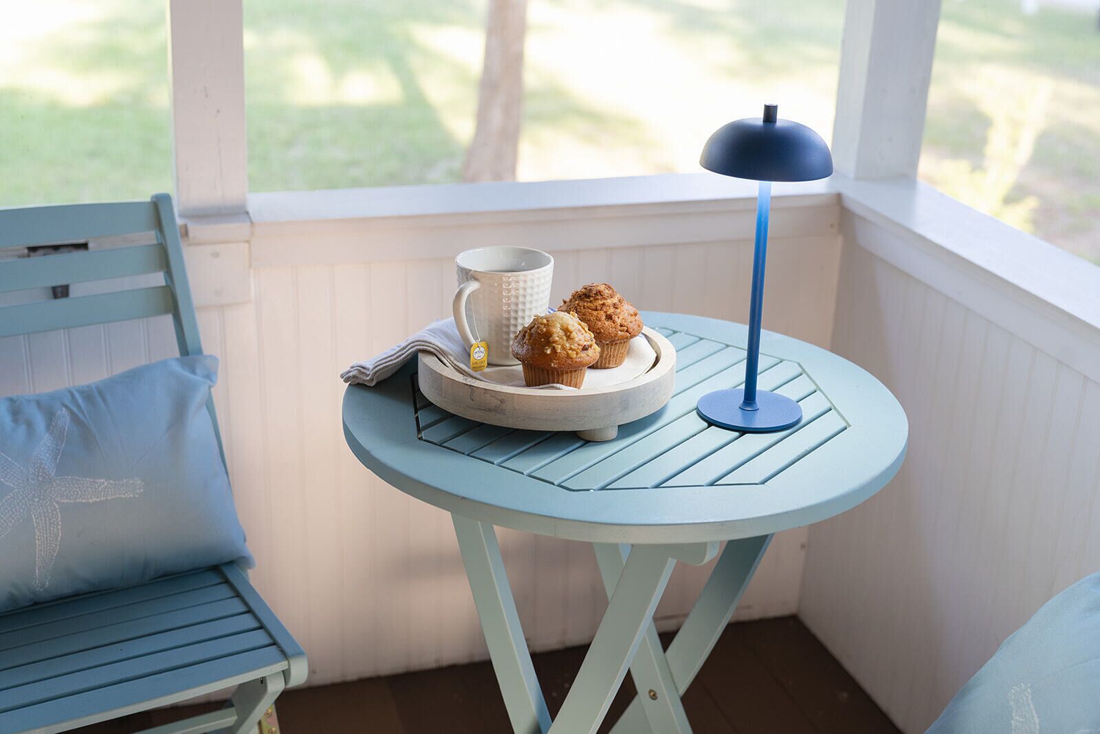 A small round table with a white mug, a plate with muffins, and a blue lamp on a porch.