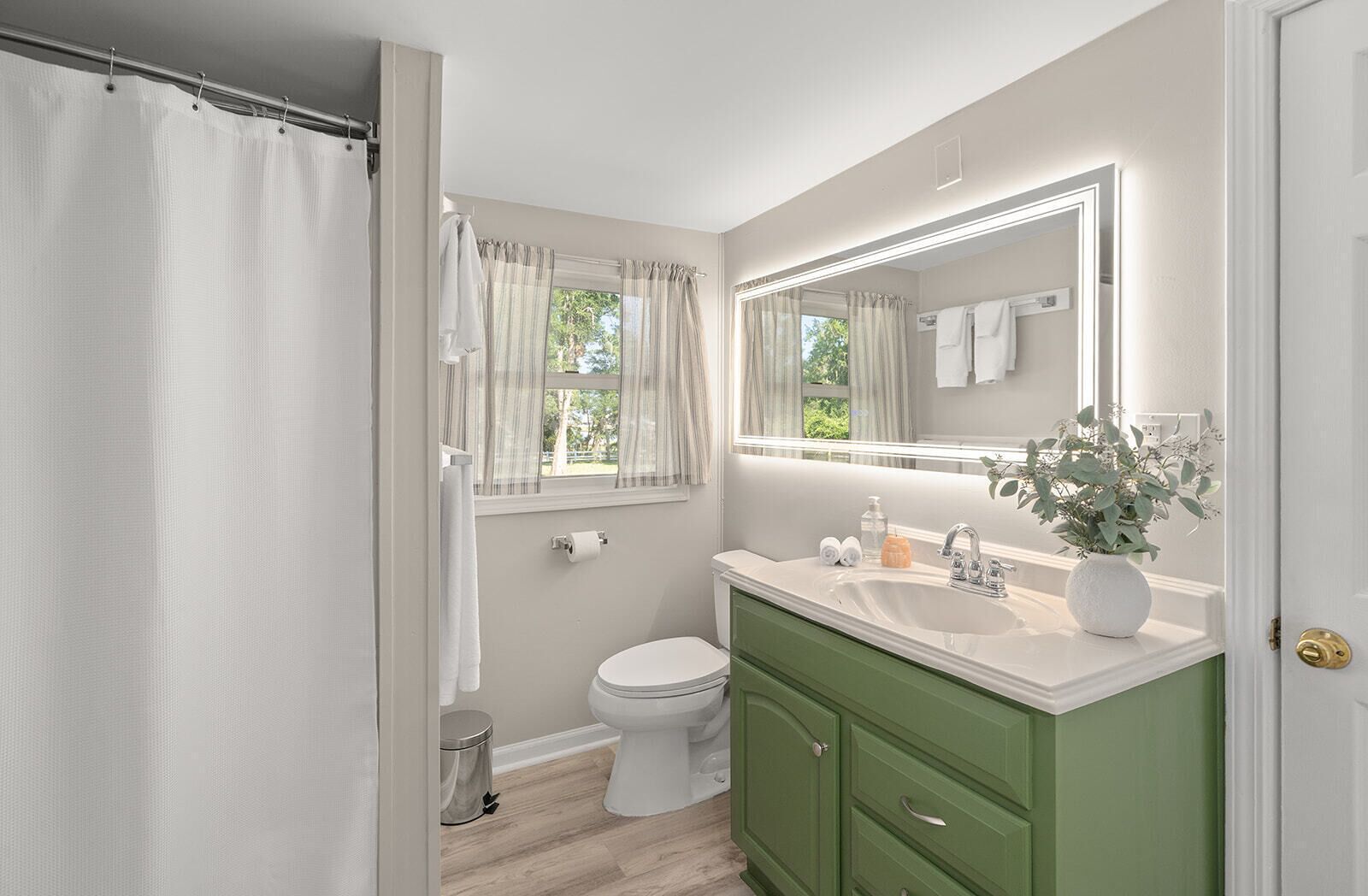A bathroom with a white toilet, a green vanity, a large mirror, and a window with curtains.