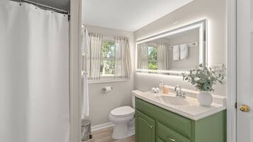 A bathroom with a white toilet, a green vanity, a large mirror, and a window with curtains.