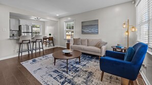 Uma sala de estar moderna com um sofá, uma poltrona azul, uma mesa de centro e um tapete. Dá para ver uma cozinha ao fundo, com uma geladeira e um espaço de jantar.