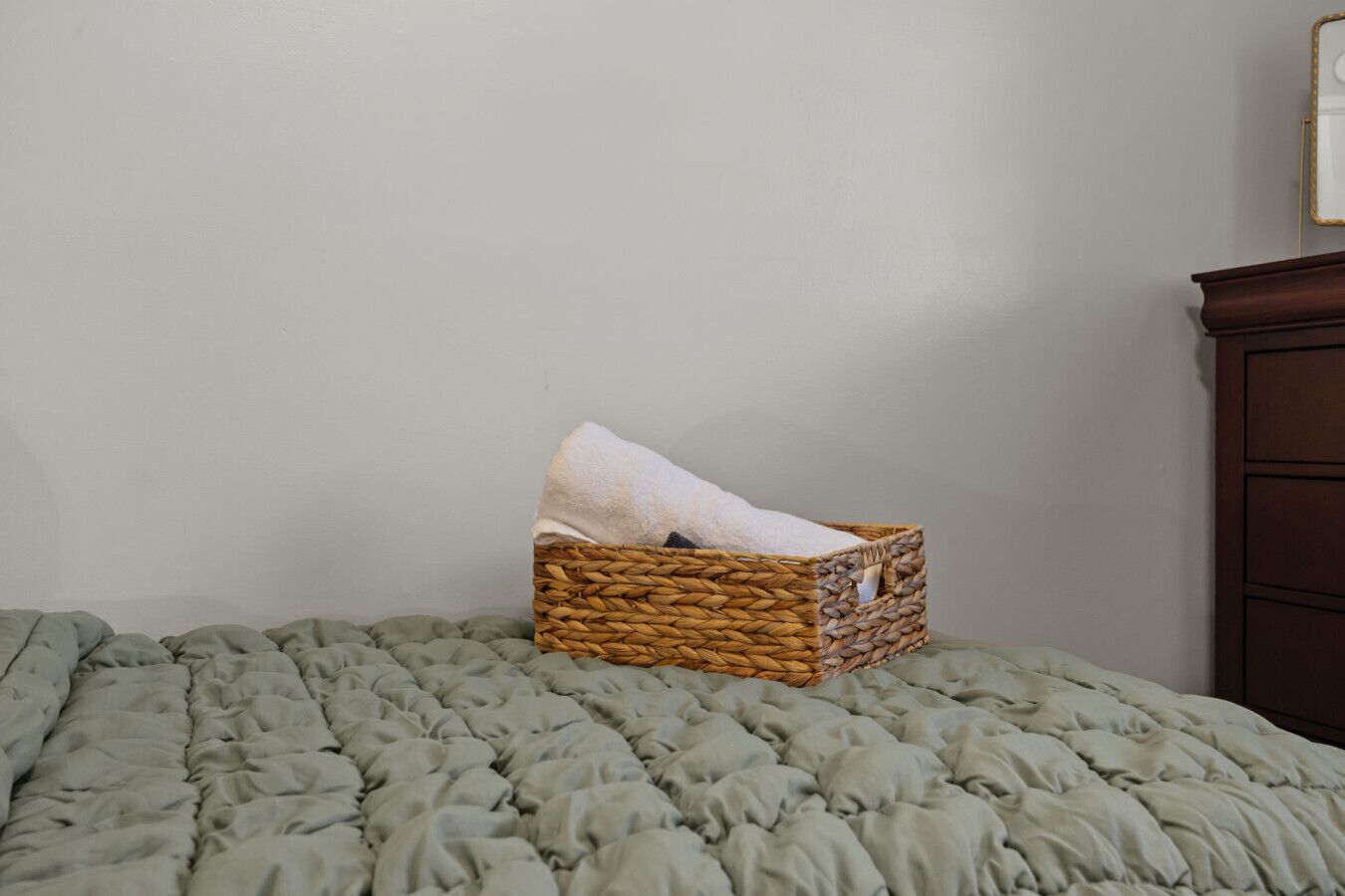 A single woven basket on a bed with a green quilt, a wooden dresser to the right, and a plain wall.