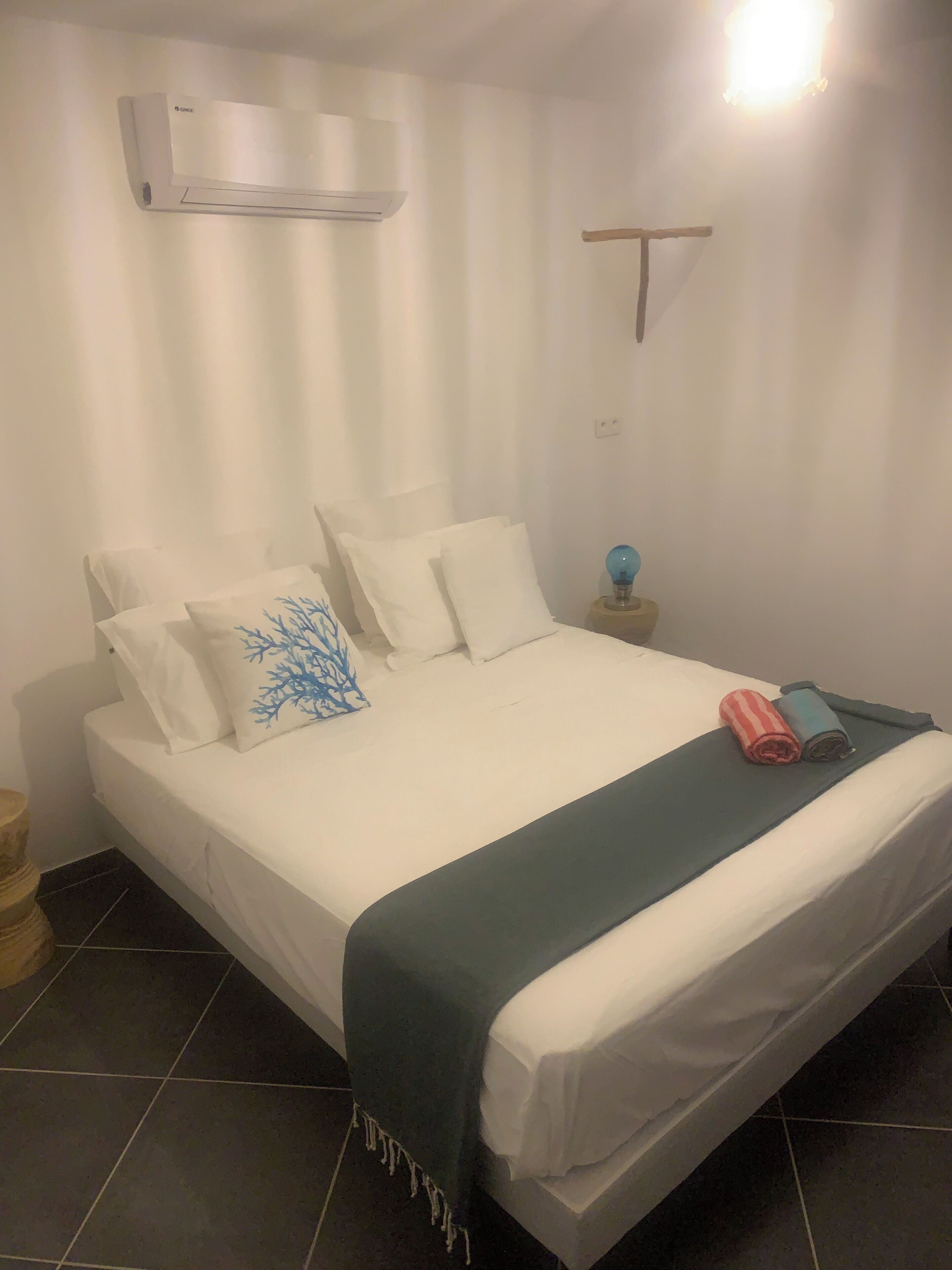 Een netjes opgemaakt bed met wit beddengoed en een blauwgroene plaid, een bedlampje en een airconditioner aan de muur.