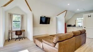 Ein Loft-Zimmer mit braunem Ledersofa, einem Schreibtisch mit Stuhl, einem an der Wand befestigten Fernseher und einer Küchenzeile im Hintergrund.