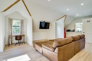 Un dormitorio tipo loft con un sofá de cuero marrón, un escritorio con silla, un televisor montado en la pared y una zona de cocina al fondo.
