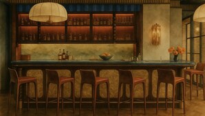 A bar with a long counter, multiple stools, and shelves with bottles. - Hotel Daphne (Houston)