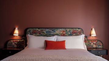 Uma cama com almofadas brancas e vermelhas, cabeceira florida e duas luminárias laterais.
