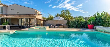 A swimming pool with a patio area, a covered seating area, and a house with a tiled roof.