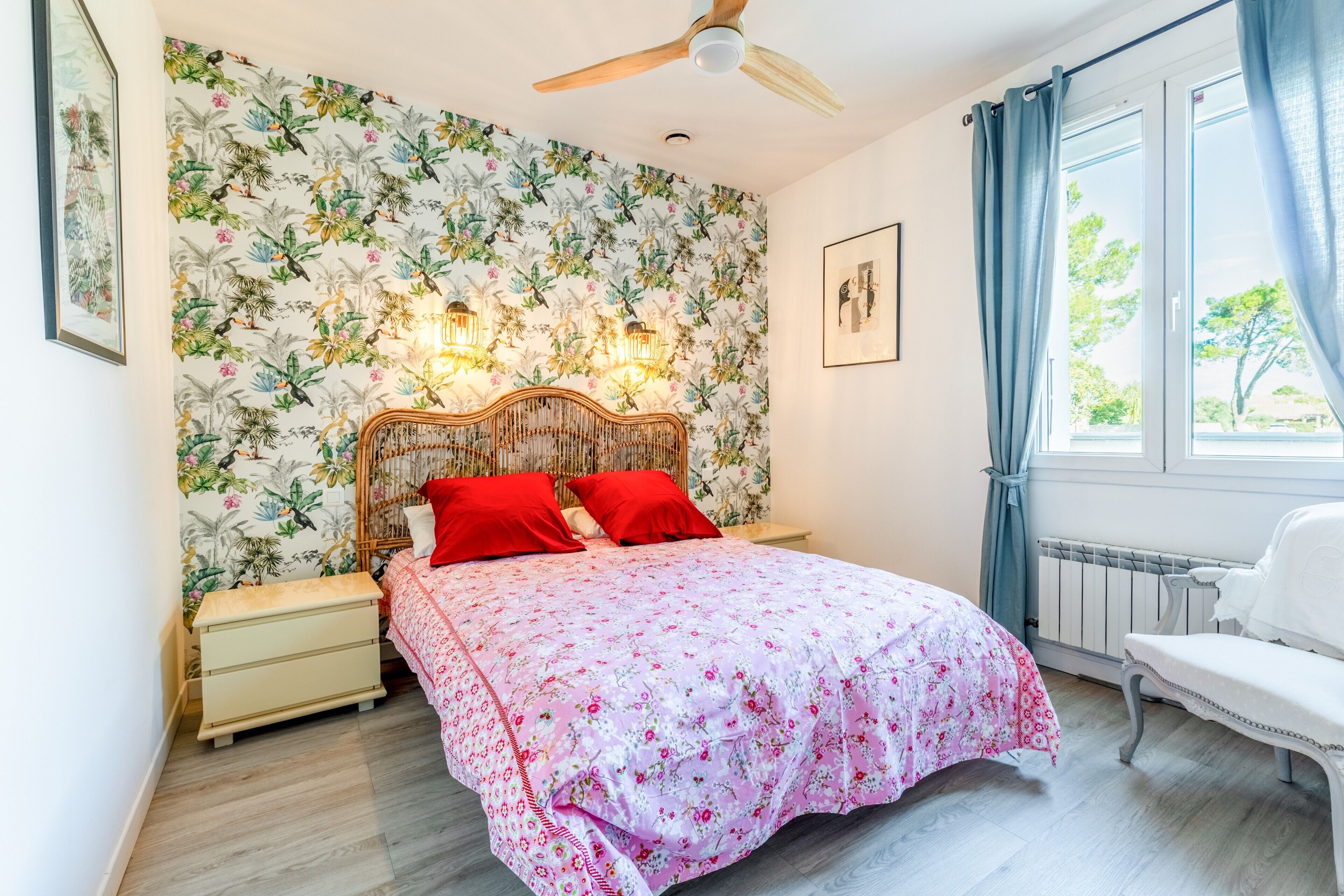 Ett sovrum med en blommig tapet, en säng med röda kuddar, ett träfärgat sänggavel, sängbord, en vit stol och en takfläkt.