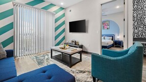Ein modernes Wohnzimmer mit einer Couch, einem Couchtisch und einem Fernseher. Eine Schiebetür mit Jalousien, eine strukturierte Wand und ein Bett im angrenzenden Raum.